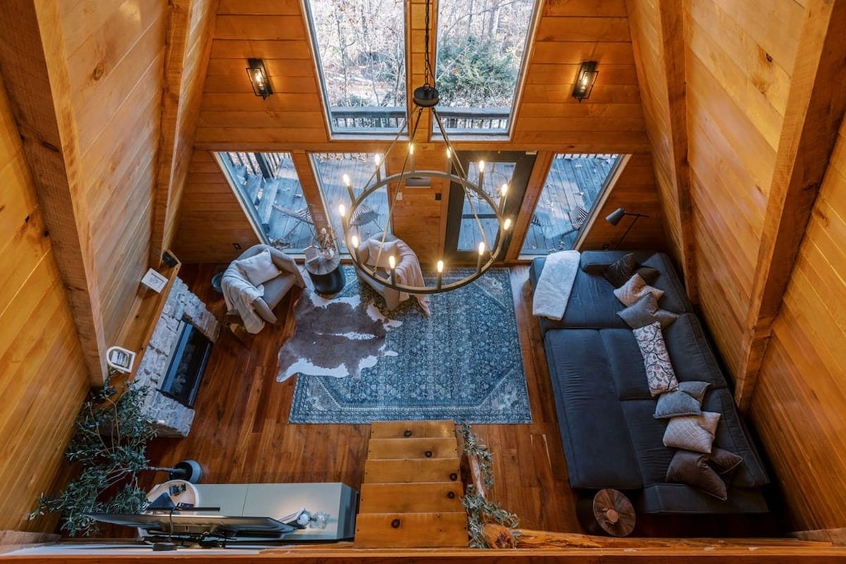 An aerial view captures the open living space of the cabin, featuring a sectional sofa adorned with decorative pillows. A circular chandelier hangs above, illuminating the wooden interiors. Large glass doors invite natural light, leading to an outdoor deck surrounded by trees. A plush area rug adds comfort.