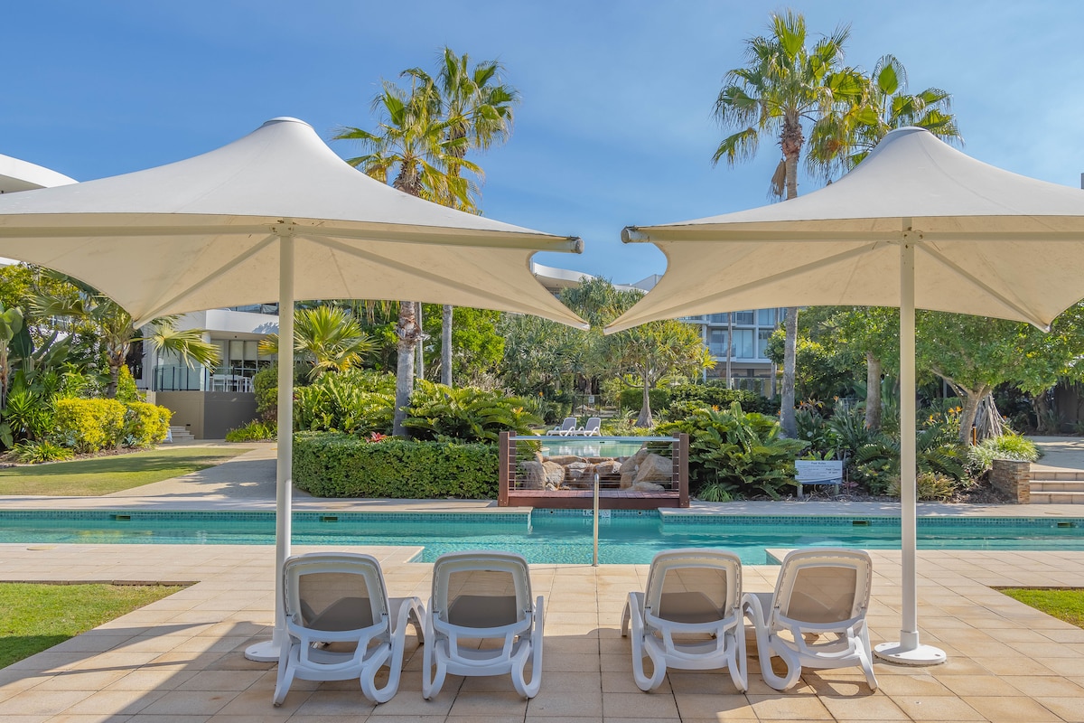A serene view of the resort's lagoon pool is captured, framed by two large umbrellas providing shade. Lounge chairs are arranged in a row on the tiled deck, overlooking lush tropical gardens and palm trees, enhancing the tranquil setting.