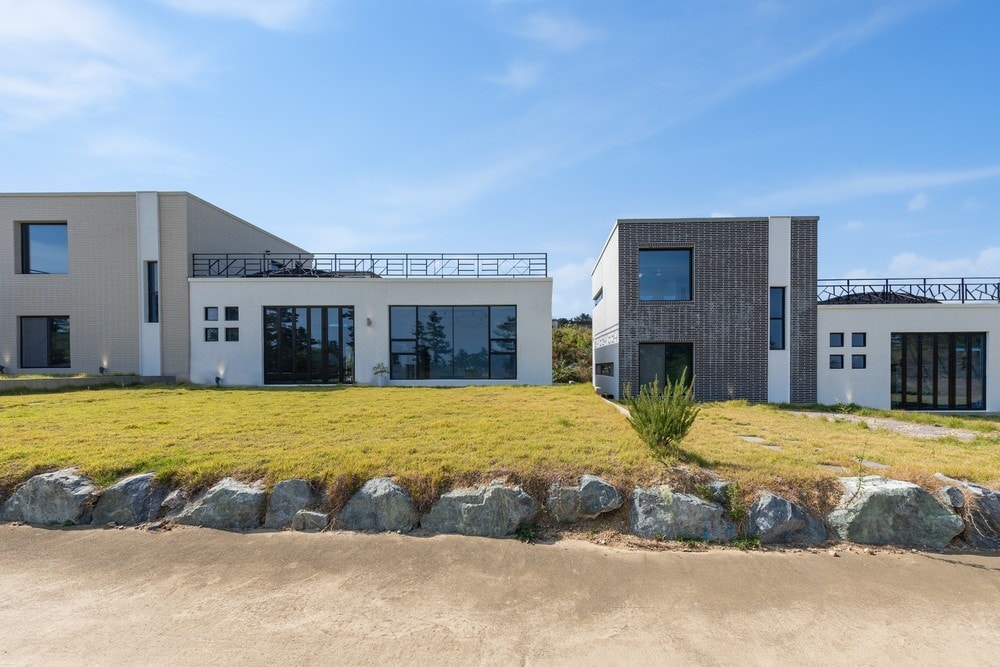 Two contemporary buildings with large glass windows are set against a clear blue sky. The structures are surrounded by a well-maintained grassy area, with natural stone borders lining the path. A spacious terrace is visible on the top level, providing outdoor access.
