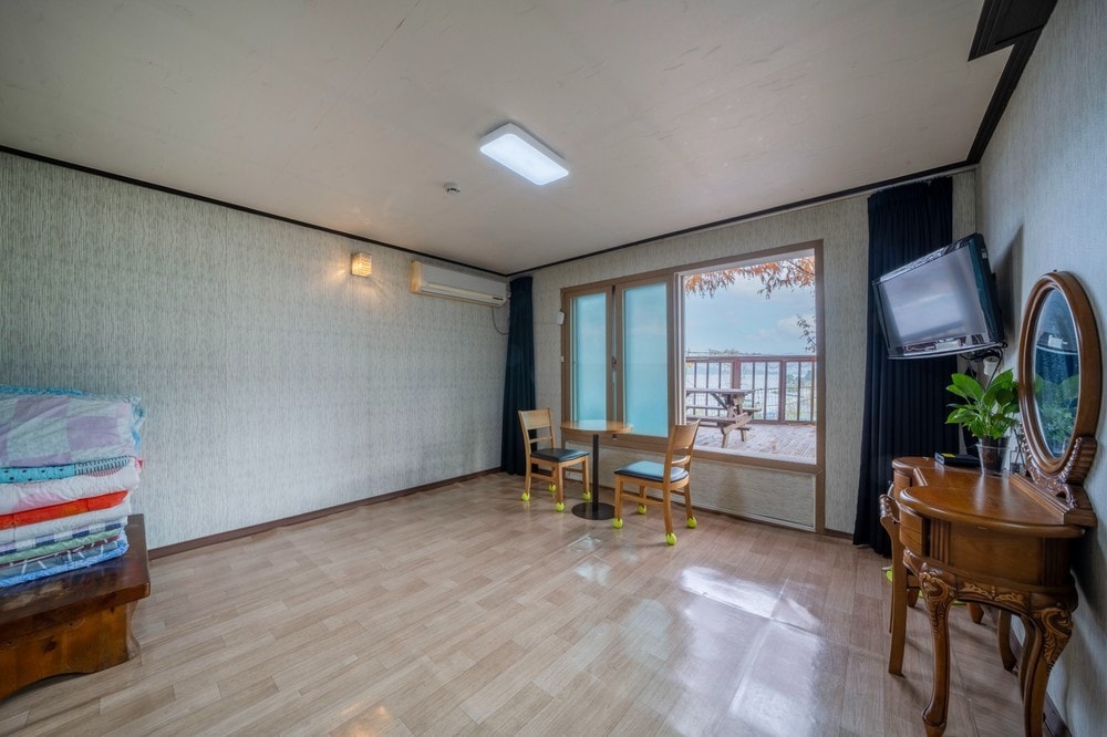 The spacious room features a wooden table and two chairs, with a large window offering views of the outdoor area. A stack of colorful bedding is displayed nearby. A flat-screen TV and decorative mirror complete the functional space.