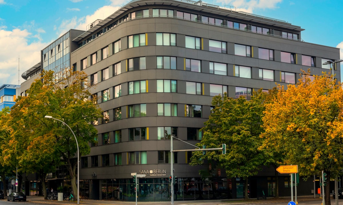 A modern, multi-story building features a sleek facade with large windows reflecting a clear sky. Surrounding trees exhibit autumn foliage, adding a splash of color to the scene. The entrance is marked by the hotel's name, positioned prominently at the ground level.