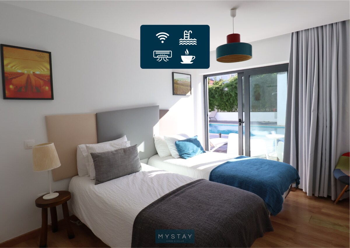 A bright room features two beds covered with light blankets, each complemented by decorative throw pillows. A bedside lamp stands next to a wooden table, and a large window offers views of the swimming pool and outdoor area, allowing natural light to fill the space.