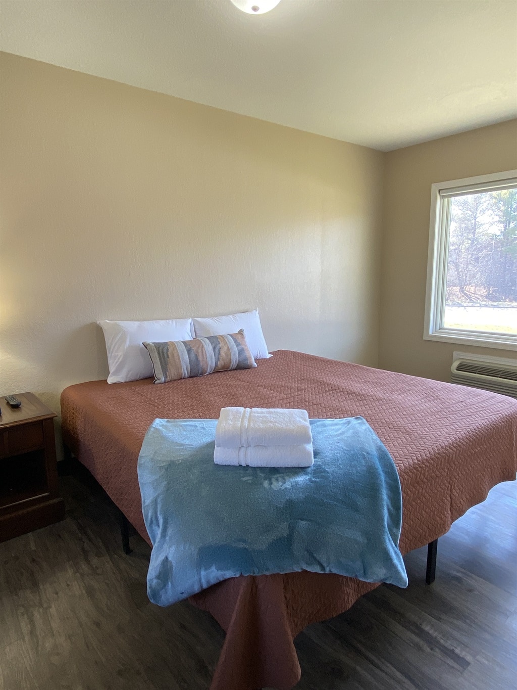 A comfortable sleeping area is depicted, featuring a spacious bed with a rust-colored coverlet and decorative pillows. A light blue towel is neatly placed on the bed. Natural light filters through a nearby window, illuminating the warm-toned walls.