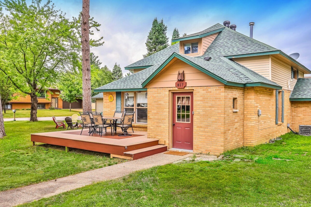 The exterior of a charming two-story cottage is showcased, featuring a spacious wooden deck and outdoor seating area. Large windows invite natural light into the home. A well-maintained lawn and trees provide a serene setting, with a clear pathway leading to the entrance.
