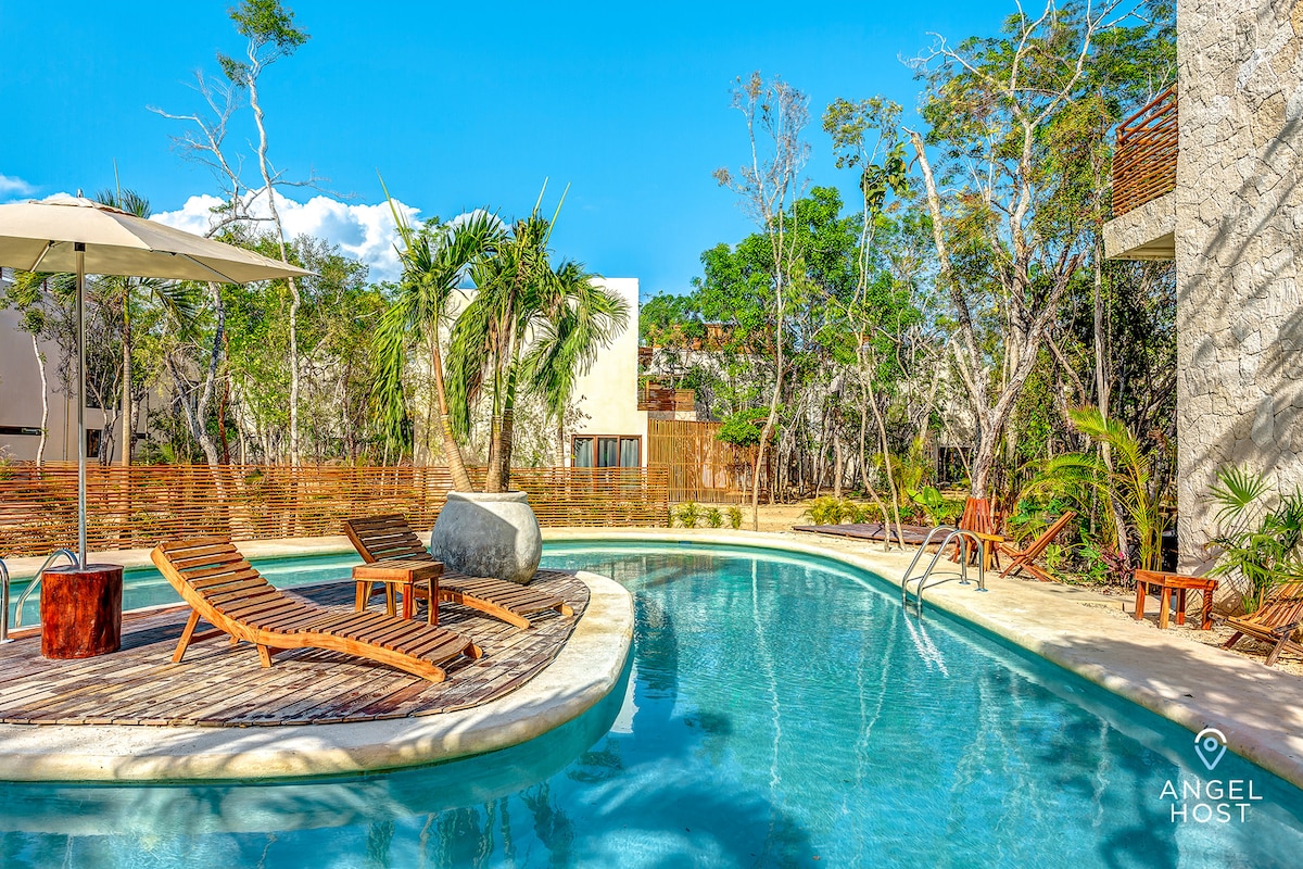 A serene pool area is surrounded by lush greenery and tropical trees. Sun loungers are arranged around the pool's edge, along with a small wooden table. An umbrella provides shade over a single lounge chair, inviting relaxation in a tranquil outdoor setting.