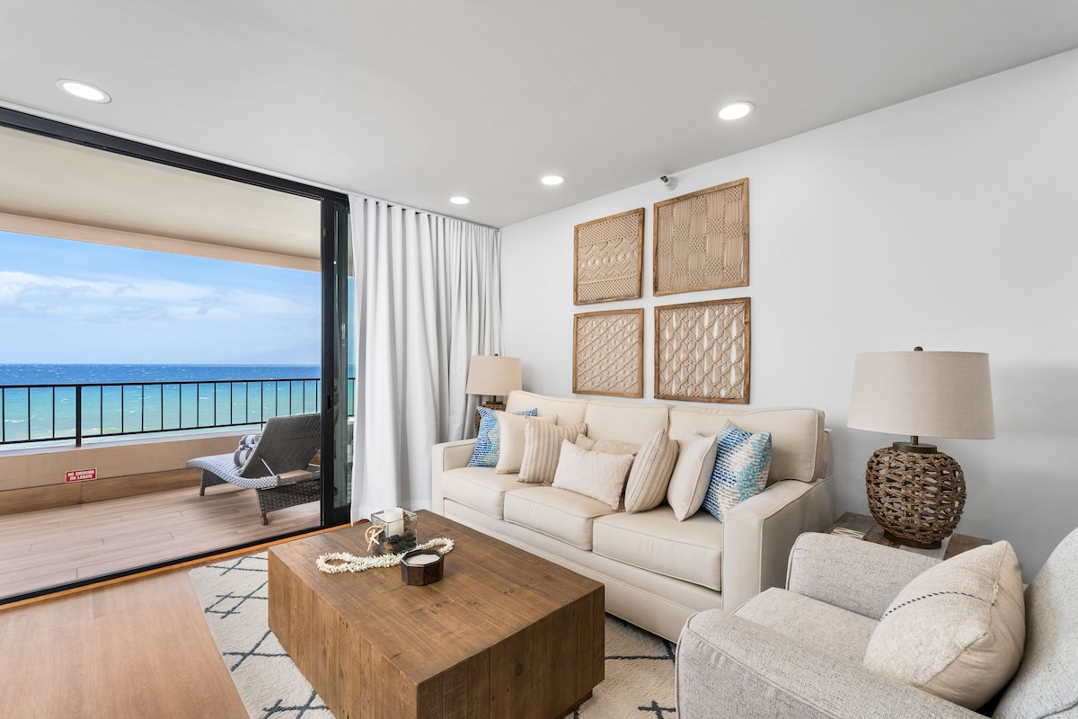 A bright and airy living room features a light-colored sofa adorned with decorative pillows. A wooden coffee table sits at the center, while two lamps provide soft lighting. Large sliding glass doors open to a private lanai with ocean views, enhancing the serene atmosphere.