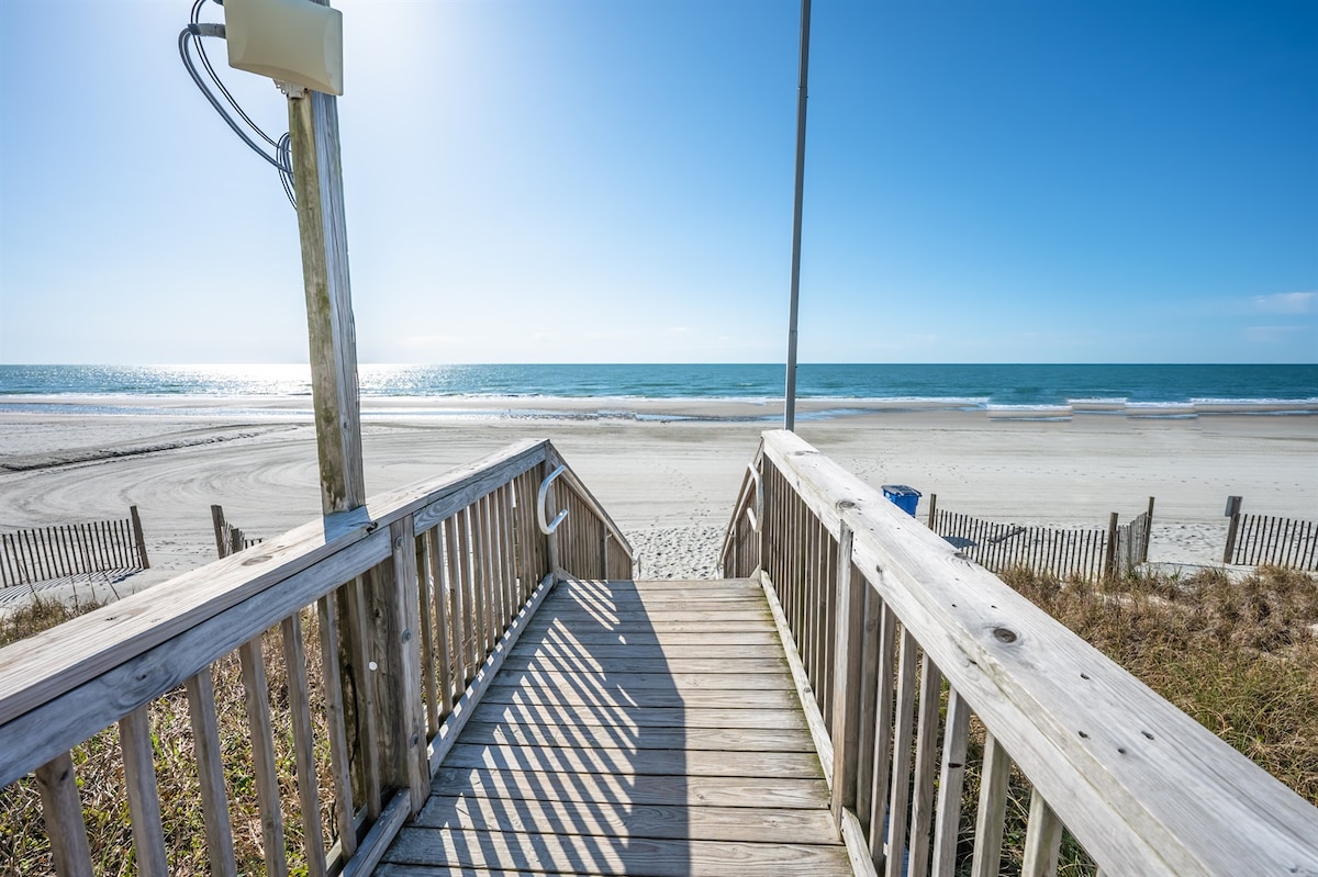 A wooden boardwalk leads down to the sandy beach, with direct views of the ocean. The calm waters reflect the sunlight, creating a serene atmosphere. Fences and grassy dunes frame the pathway, emphasizing the proximity to the shoreline.