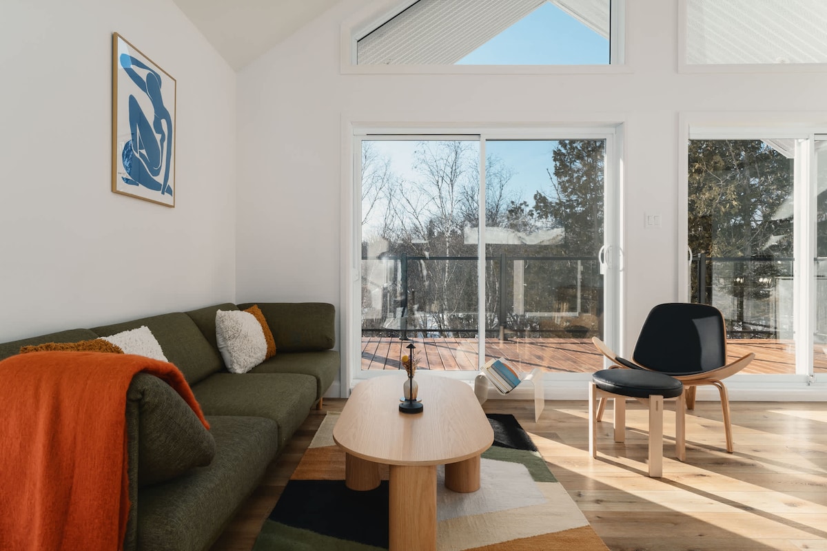 A bright living room features a green sectional sofa adorned with a textured throw. A wooden coffee table sits in front, accompanied by a modern chair. Large glass sliding doors open to a spacious deck, framed by a tree-lined view and natural light streaming in.