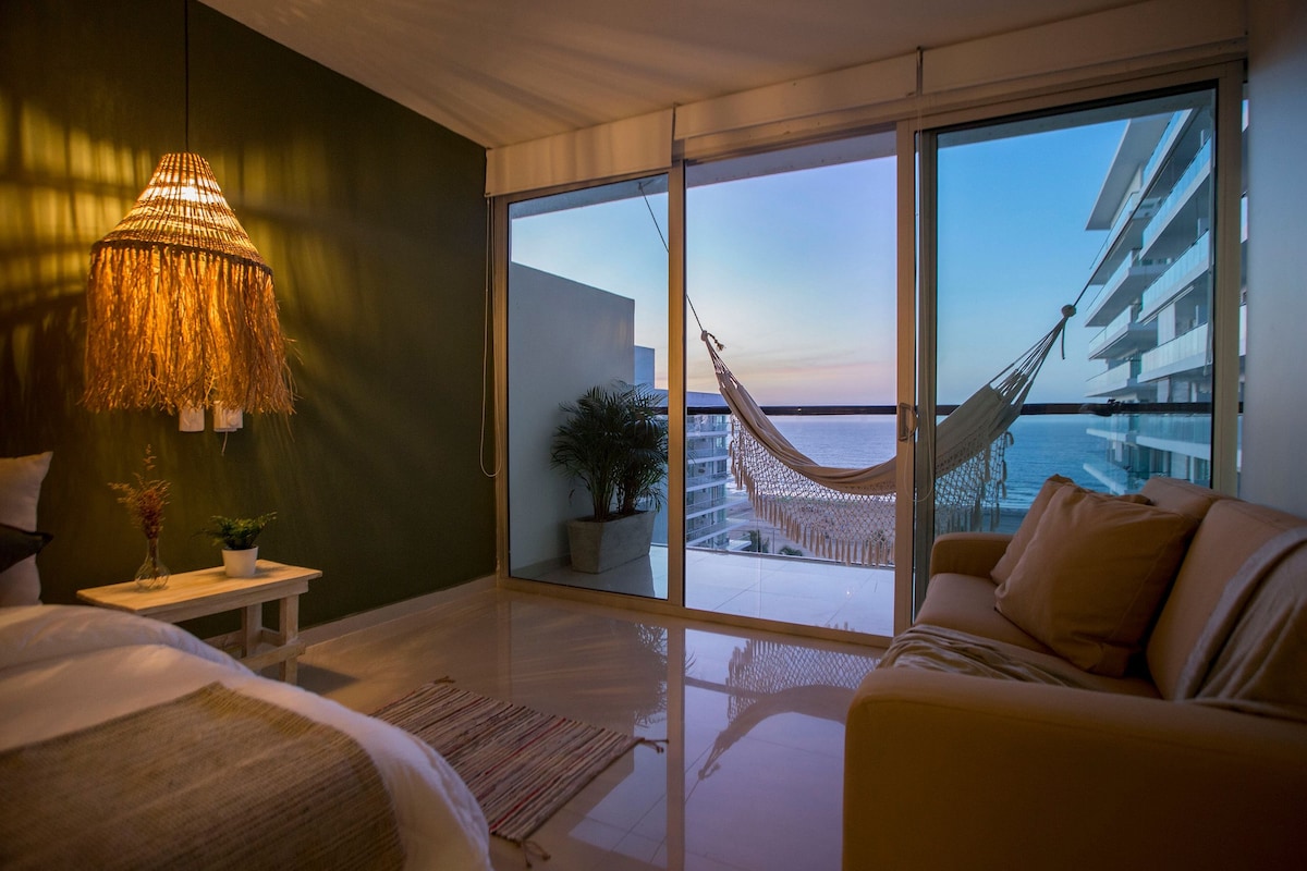 A cozy corner of a room is highlighted by a warm light fixture and a comfortable sofa. Large glass doors offer a view of the ocean and a hammock is positioned on the balcony, allowing for relaxation while enjoying the sea breeze.
