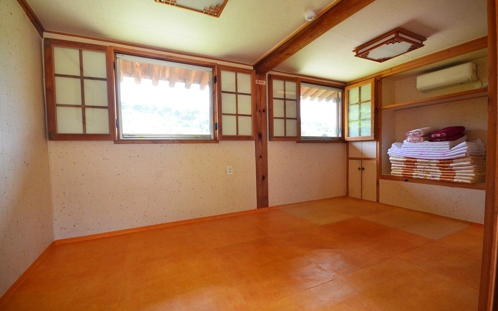 A light and airy room features two large windows with wooden frames, providing natural light. The orange flooring complements the neutral walls. A shelf is neatly arranged with stacked towels, and an air conditioning unit is installed to enhance comfort.