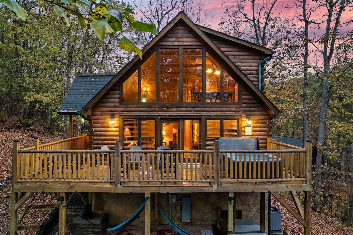 A spacious cabin is showcased, surrounded by trees and bathed in warm light during sunset. Large glass windows provide panoramic views of the forest. An inviting deck features wooden railings, outdoor furniture, and hammocks, emphasizing the outdoor relaxation space.