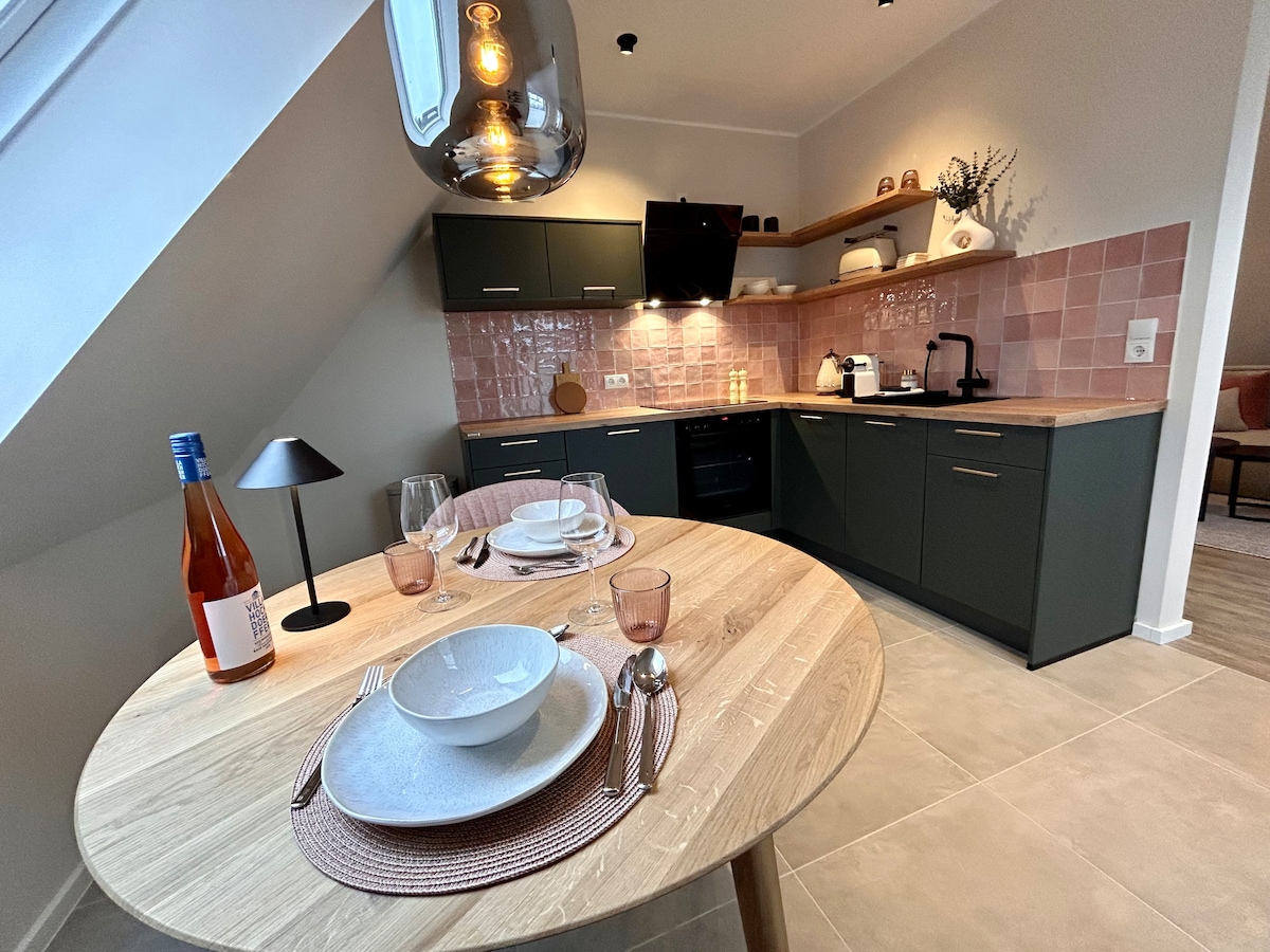 A modern kitchen is highlighted by an elegantly set wooden dining table. Soft pink tiles create a warm backdrop behind the dark cabinetry. Under-cabinet lighting features a sleek, contemporary design, while stylish dinnerware and glassware are thoughtfully arranged. A bottle of wine adds a subtle touch.