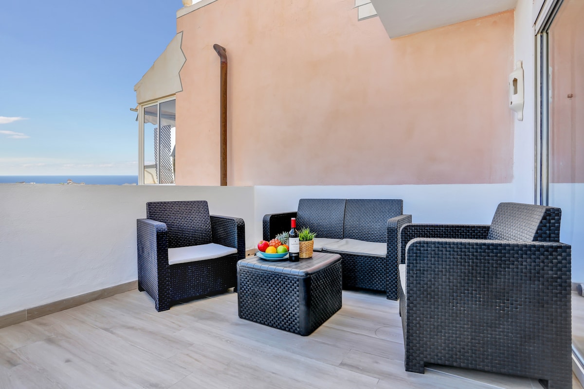 A furnished terrace is presented with stylish outdoor seating, featuring two armchairs and a coffee table. A bowl of fresh fruit decorates the center of the table, and expansive views of the ocean can be seen in the background, complemented by clear blue skies.