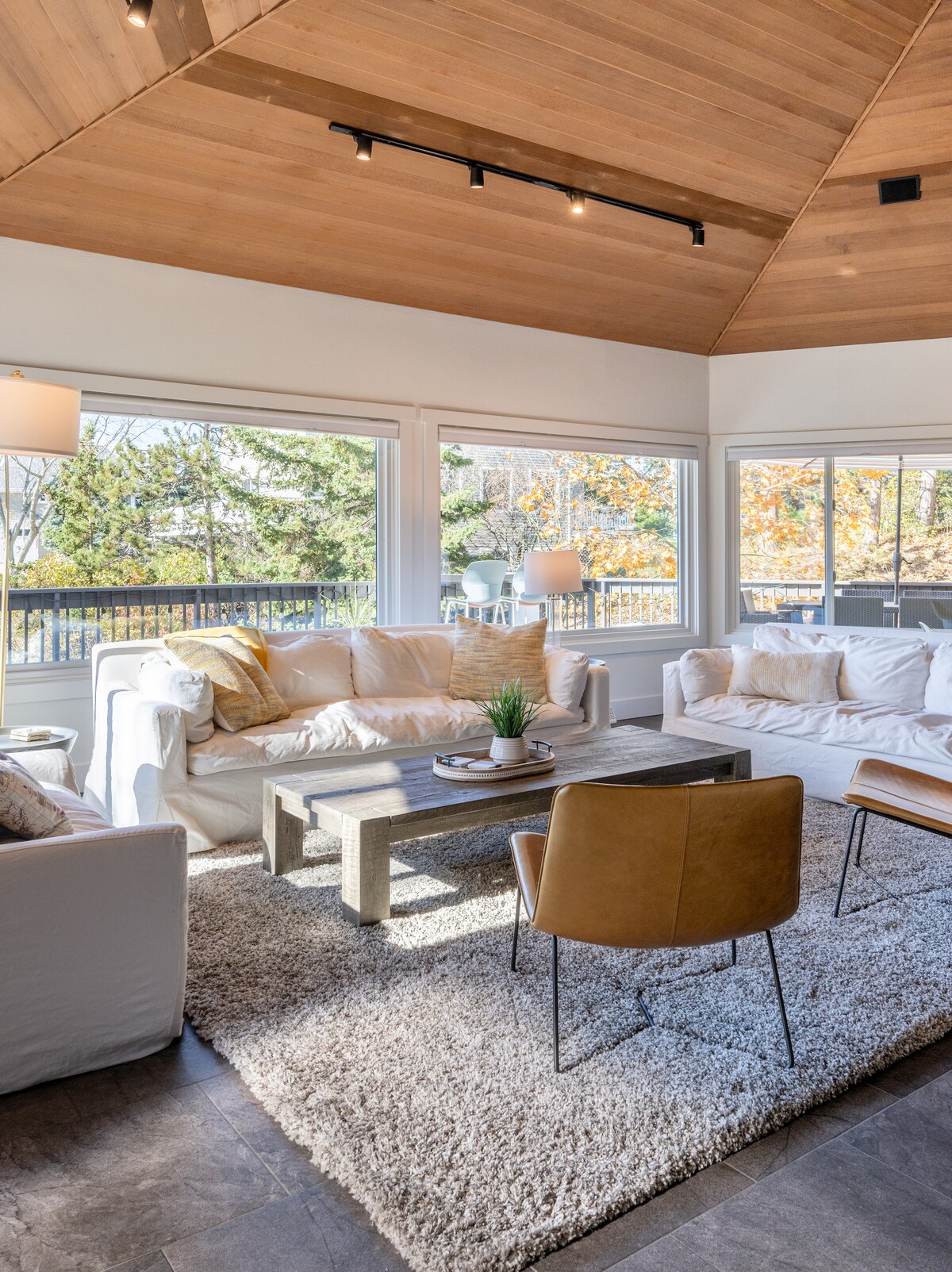 A bright and airy living area features a high wooden ceiling and large windows showcasing natural light and scenic views. Soft, oversized sofas and a plush rug create a comfortable seating arrangement around a rustic coffee table, complemented by a contemporary accent chair.
