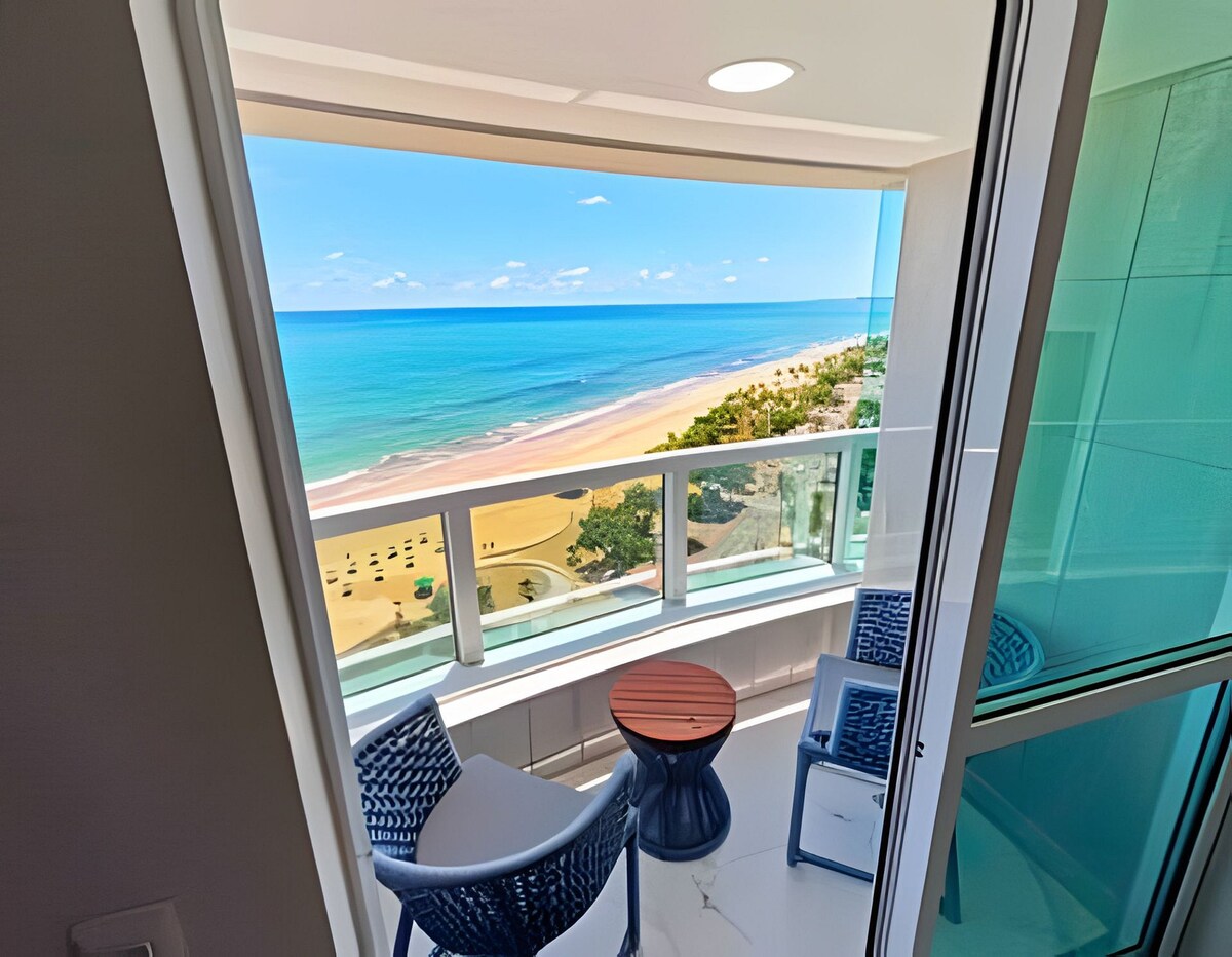 A balcony area offers stunning ocean views, showcasing a sandy beach and vibrant blue waters. Two comfortable chairs are positioned around a round table, providing a perfect spot for relaxation while enjoying the scenery. Natural light floods the space through large glass doors.