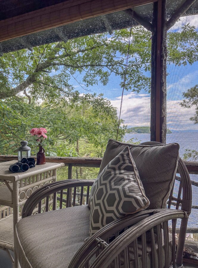 A cozy seating area is presented on a screened porch, featuring a woven chair with a decorative cushion. A small table holds a vase with fresh flowers and binoculars, offering a view of lush greenery and the tranquil lake beyond.