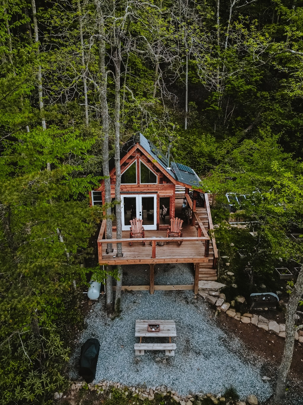 An off-grid treehouse is nestled among tall trees, showcasing a wooden deck with two chairs positioned for relaxation. Solar panels adorn the slanted roof, and a stone pathway leads to a picnic area nearby, set on a bed of gravel surrounded by lush greenery.