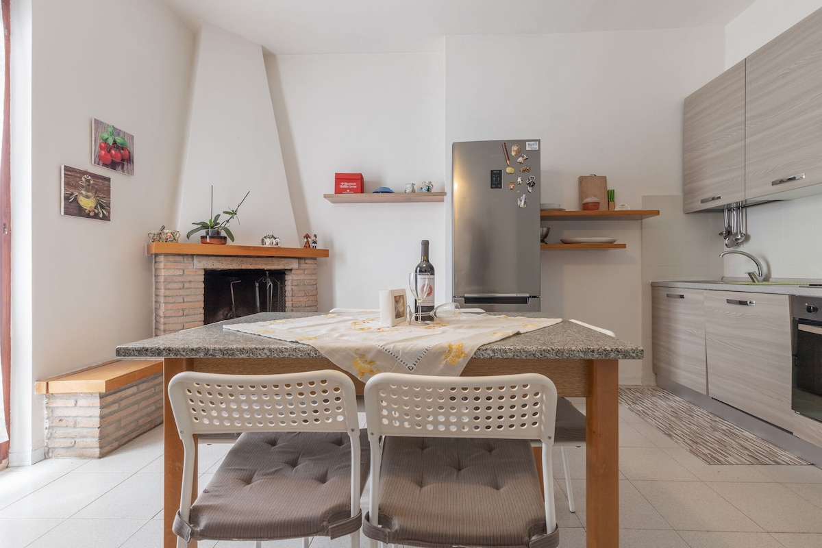 A well-equipped kitchen features a modern design, including a countertop with a grey granite surface and two white chairs. The kitchen extends to a functional area with a silver refrigerator and wood shelves. A brick fireplace adds a warm touch to the space.