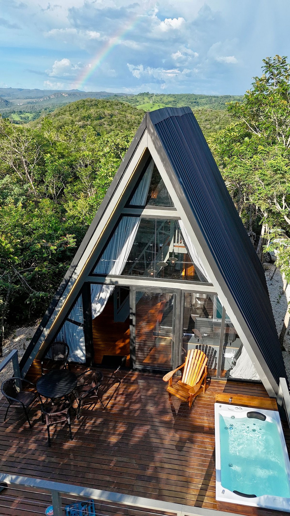 An A-frame structure is set against a backdrop of rolling hills and greenery, featuring large glass doors that invite natural light. A private deck holds seating arrangements and a jacuzzi, with a rainbow arching gracefully in the sky above.
