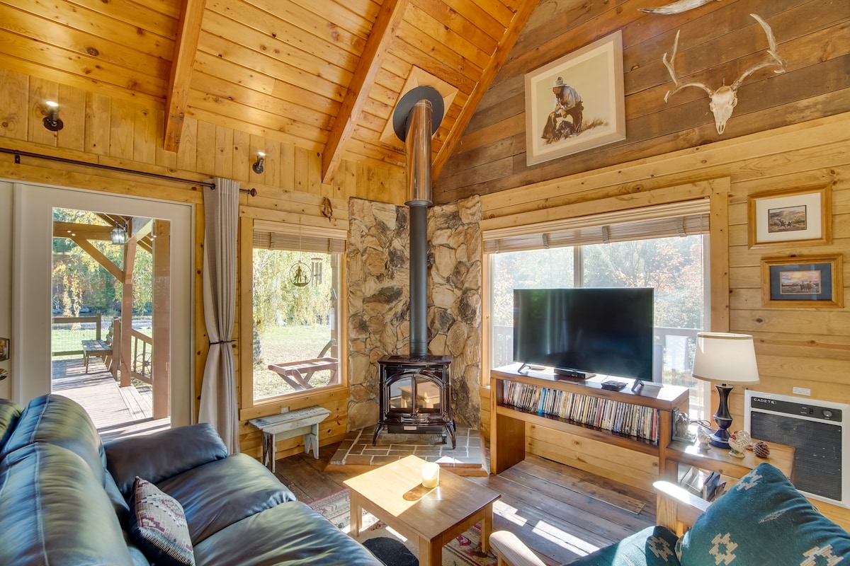 The living area showcases vaulted wooden ceilings with exposed beams. A stone fireplace stands as a focal point, accompanied by a dark leather sofa and wooden coffee table. Light filters through large windows, illuminating a Smart TV and decorative wall art.