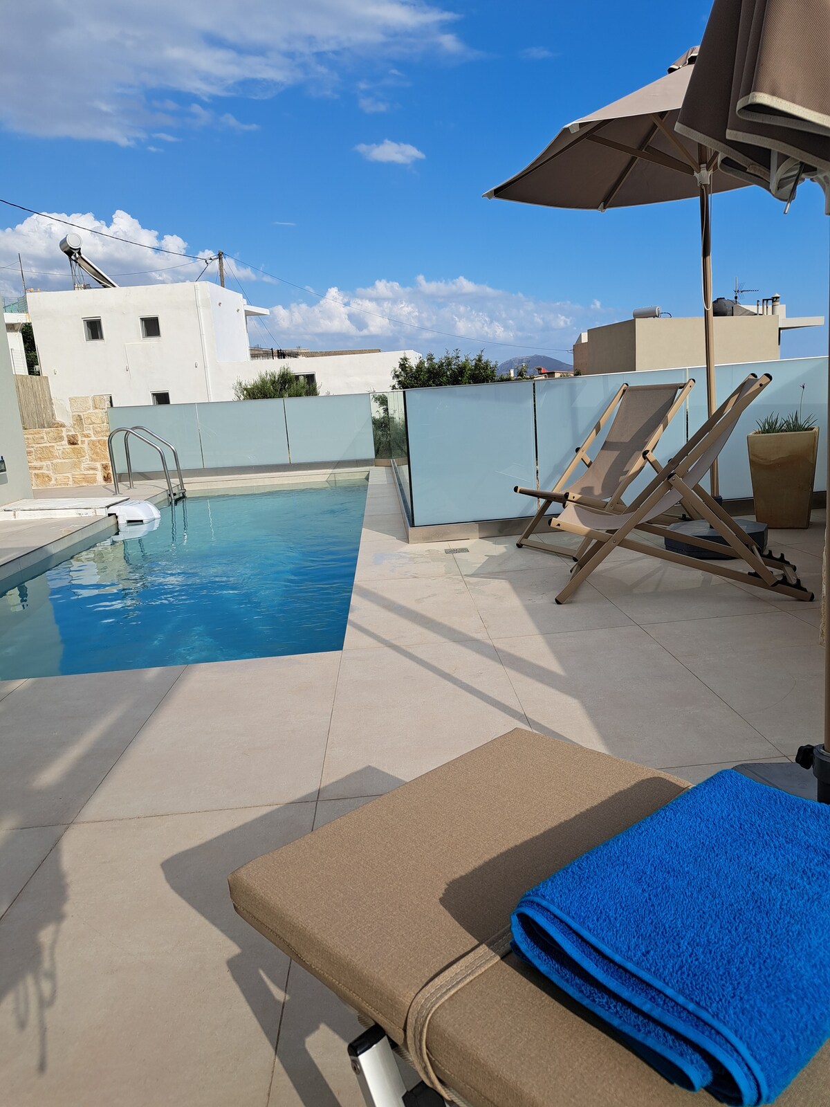 A private pool is featured, surrounded by light-colored tiles and glass panels that enhance openness. Two wooden lounge chairs are positioned nearby, with a blue towel placed on one of the chairs. The sky appears clear with scattered clouds in the background.