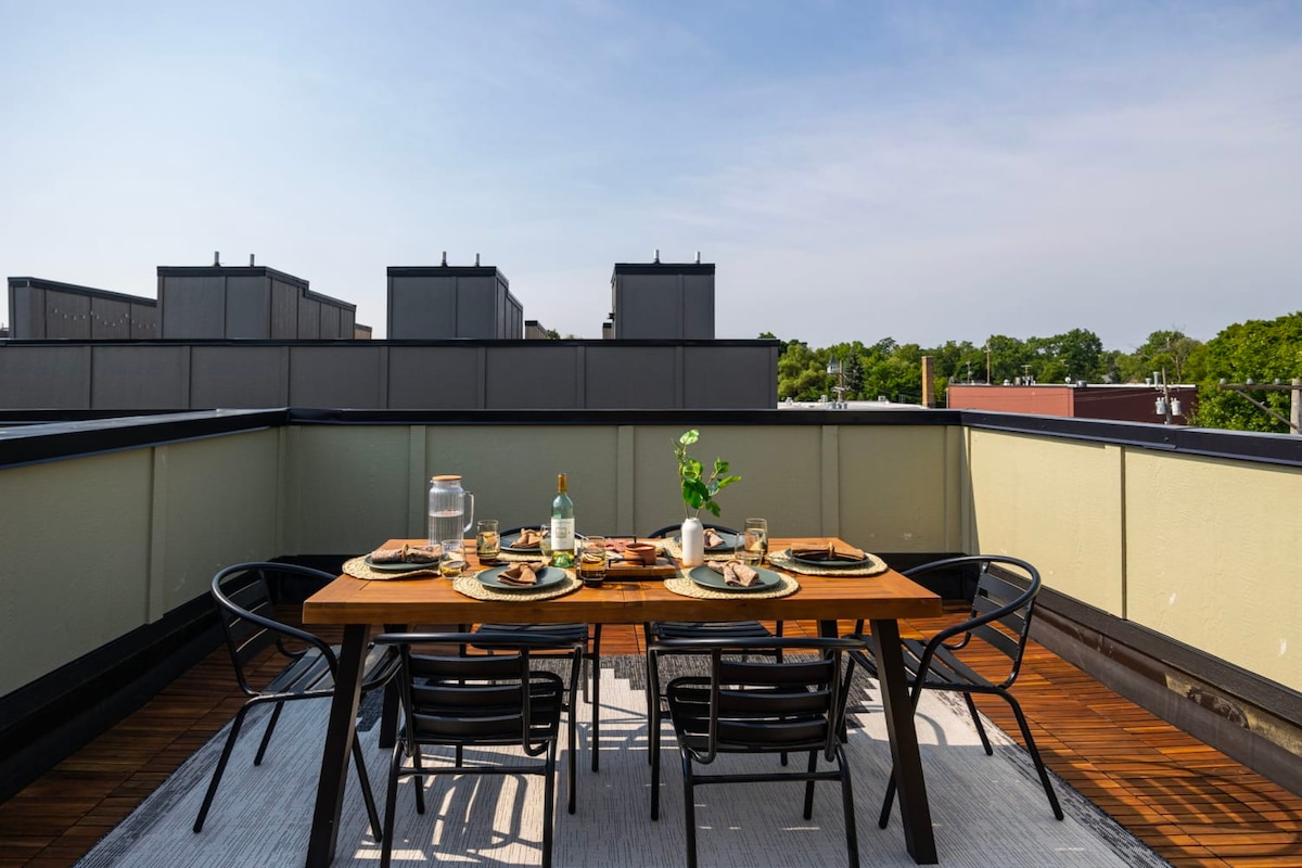 A rooftop deck is presented, featuring a wooden dining table set for six, surrounded by black chairs. A bottle of water and glasses are arranged on the table, alongside a plate of snacks. The area is enclosed with low walls, overlooking nearby rooftops and a green landscape.