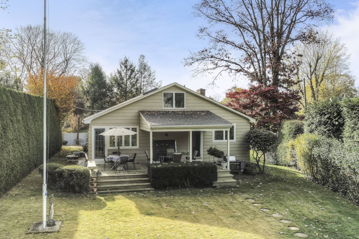 The exterior of the property features a spacious backyard with well-maintained grass and garden areas. A cozy porch is equipped with outdoor seating and an umbrella for shade. Surrounding greenery adds privacy, while steps lead to the inviting entrance of the house.