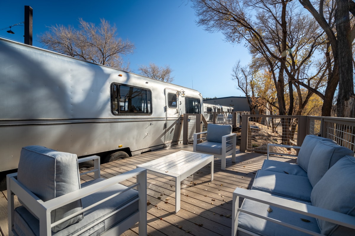 A private outdoor deck features comfortable seating arrangements with light blue cushions, accompanied by a sleek white table. The modern Avion trailer is visible nearby, set against a backdrop of clear blue skies and trees, creating an inviting outdoor space.