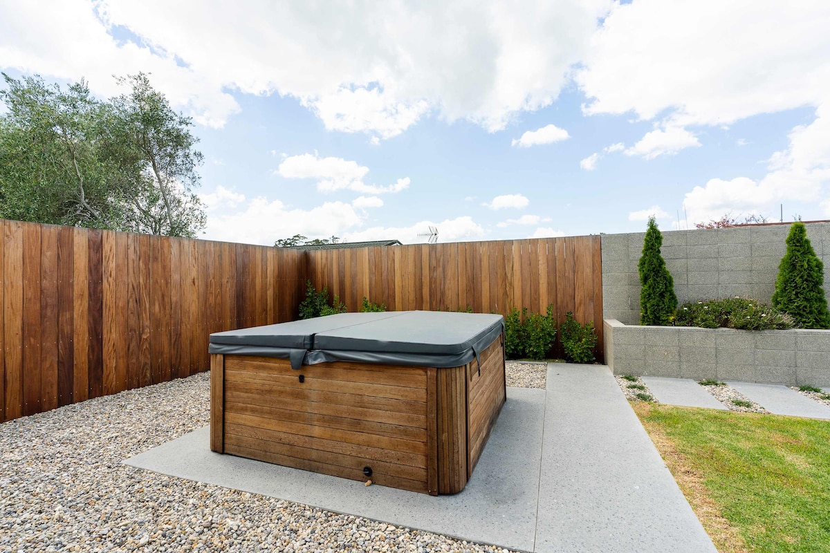 A spa pool is positioned on a stone patio, surrounded by a gravel area and tastefully landscaped borders. The wooden structure features a protective cover and is set against a backdrop of wooden fencing for privacy. Cloudy skies provide natural lighting.