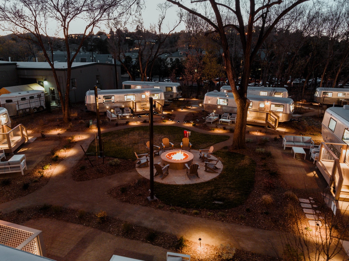 An aerial view of the property reveals multiple vintage trailers arranged around a central communal fire pit. Soft lighting illuminates the area, highlighting seating options and pathways lined with greenery, creating a welcoming outdoor space for gatherings.