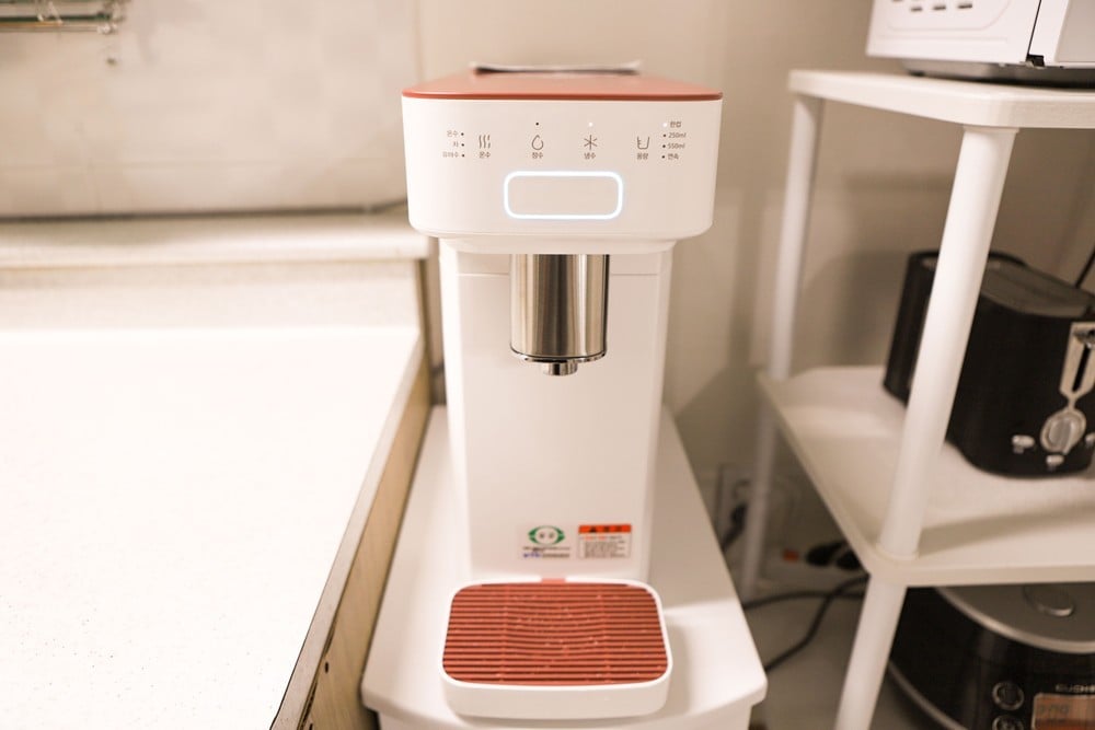 A modern beverage machine is positioned on a countertop. The machine features a sleek white design with a red accent. Controls are visible on the front, and the dispensing nozzle is at an accessible height for convenience. Surrounding appliances are neatly arranged.