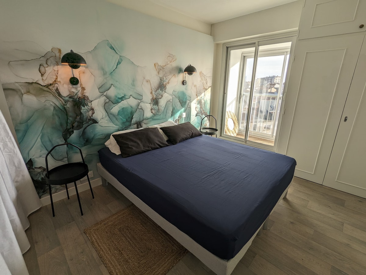 A serene bedroom features a spacious bed with a dark blue coverlet and two dark pillows. Flanking the bed are two minimalist black chairs. A decorative wall displays a soothing blend of aqua and green hues, while large bay windows allow natural light and views of the outside.