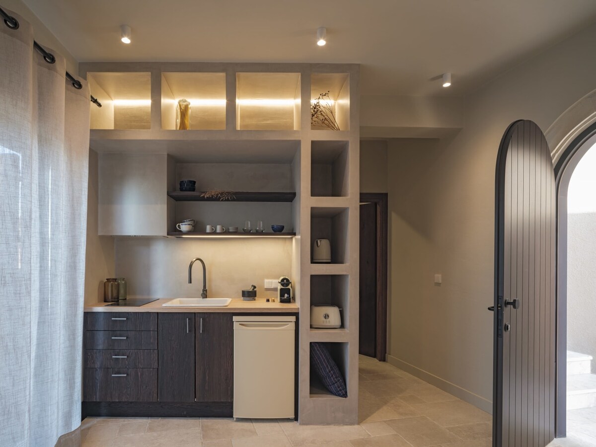 A modern kitchen area features wooden cabinetry complemented by illuminated shelving. The sink, countertop, and a compact refrigerator are visible, alongside kitchen appliances neatly arranged. Natural light filters through sheer curtains, creating a warm and inviting space.