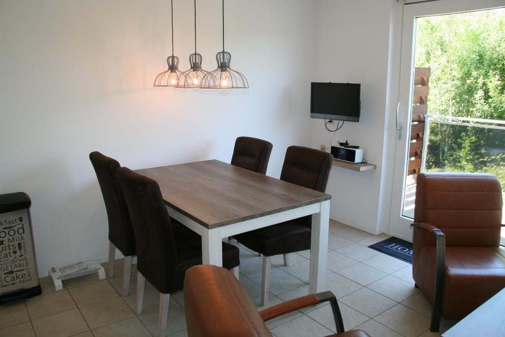 A dining area features a rectangular table surrounded by six chairs. Above the table, three pendant lights provide illumination. A wall-mounted television is visible, and a sliding door opens to a balcony. Neutral tones and light-colored tiles contribute to a bright atmosphere.