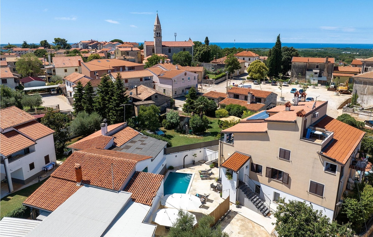 An aerial view captures the surrounding neighborhood and the shimmering pool in the enclosed property. The charming rooftops of the village are visible, along with greenery and the distant outline of the sea. The image conveys a peaceful residential atmosphere.