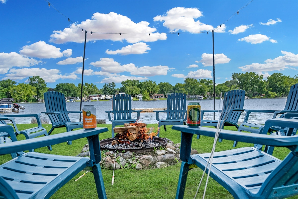 A circle of blue Adirondack chairs surrounds a firepit, with a stack of firewood at its center. The shimmering water is visible in the background, and a clear sky with scattered clouds creates a vibrant atmosphere. String lights hang above, adding to the inviting space.