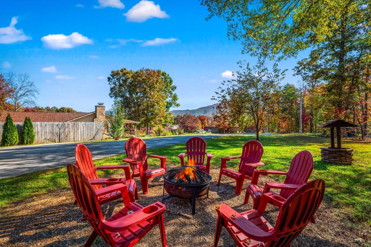 A fire pit surrounded by six red Adirondack chairs is set on a grassy area, inviting guests to gather. The serene landscape features trees displaying autumn foliage, with a view of distant mountains and a paved road in the background.