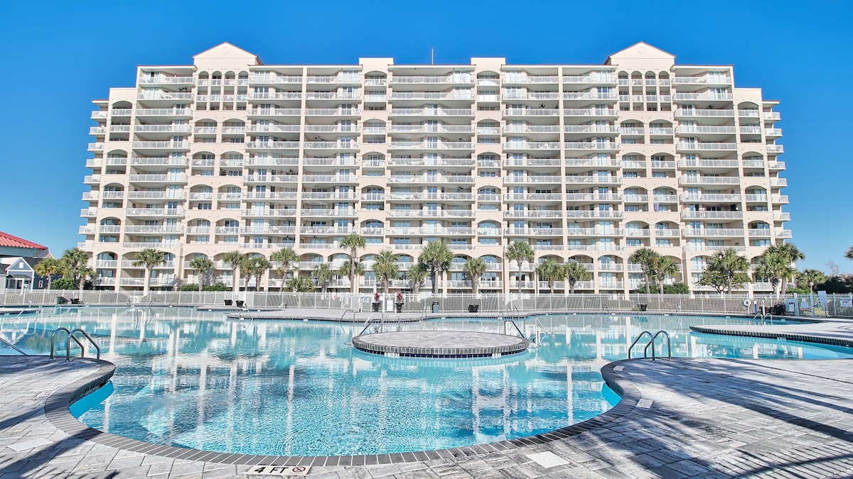 The exterior of the building is shown, featuring multiple balconies and large windows. A spacious pool area is visible in the foreground, surrounded by palm trees and lounge spaces, reflecting the clear blue sky above.