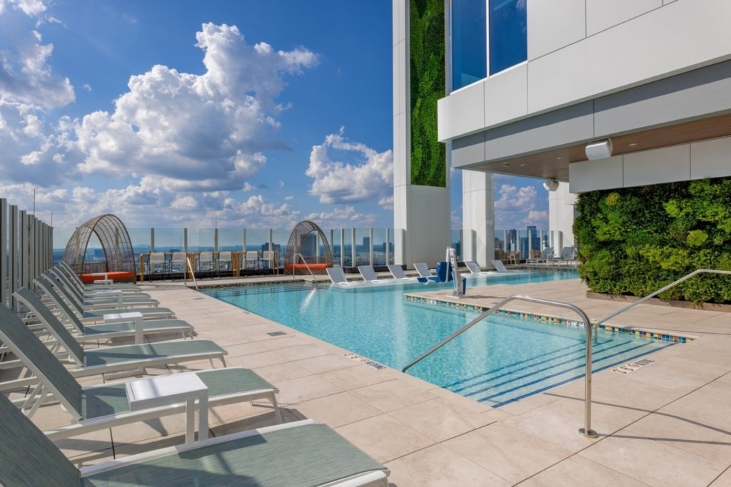 An outdoor rooftop pool area is featured, surrounded by sun loungers and elegant greenery. The clear blue water of the pool reflects the sky, complemented by a modern building structure in the background. Scenic views of the city skyline can be seen in the distance.