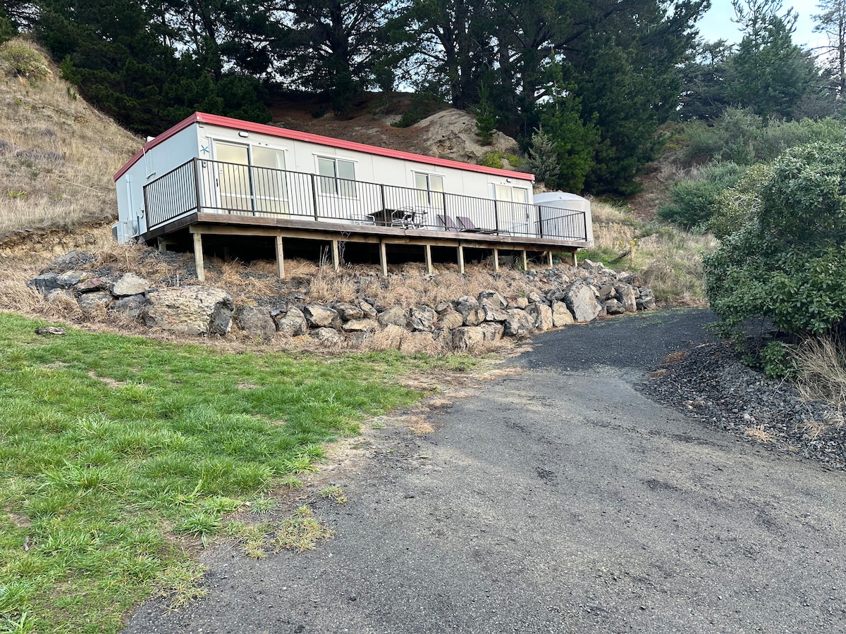 A single-story holiday unit is positioned on a hillside, featuring a raised deck with railing. The entry path leads from a rocky driveway lined with grass and shrubs, showcasing a tranquil natural setting surrounded by trees.