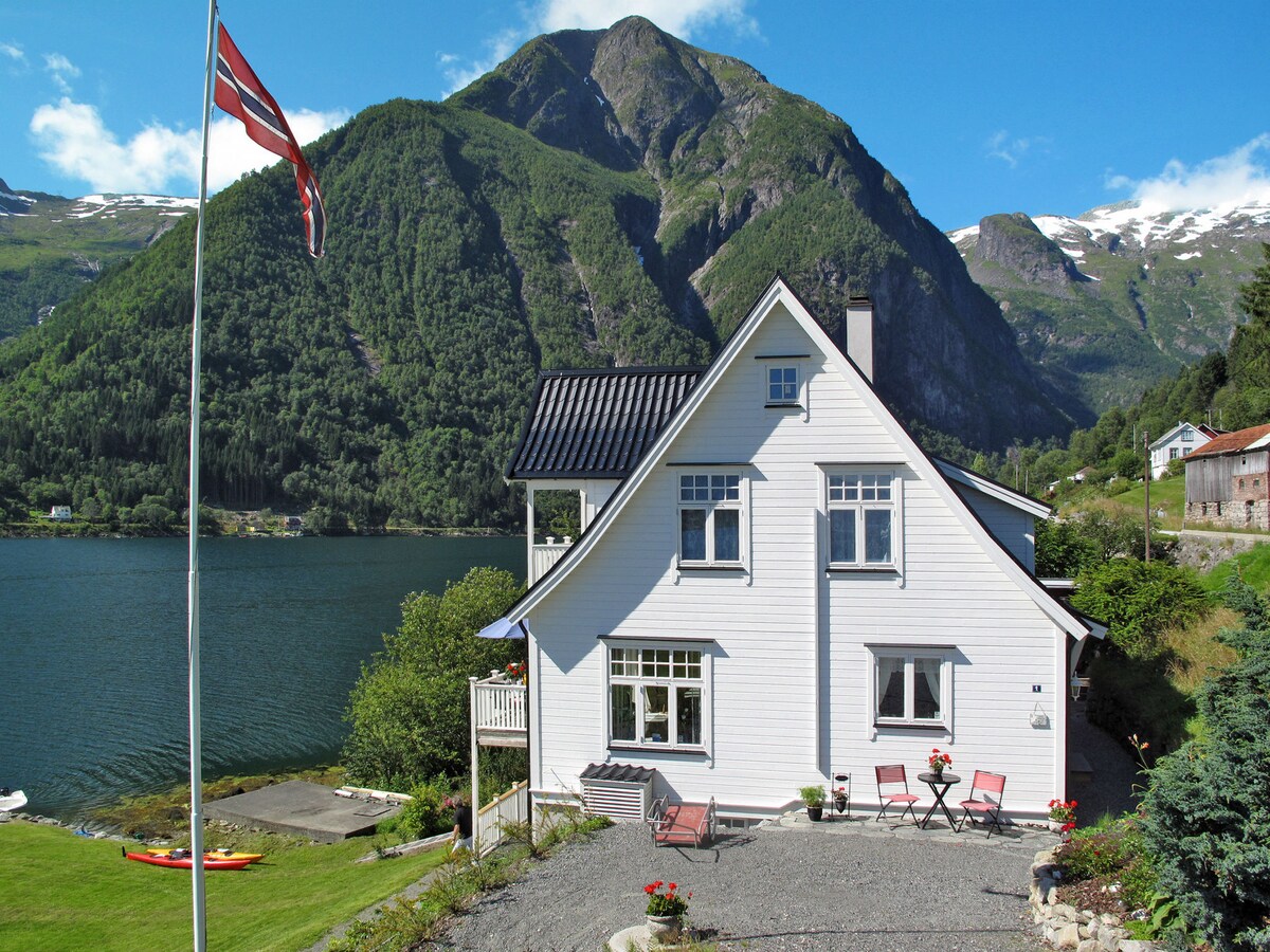 A charming white timber house is situated by the water's edge, surrounded by lush mountains. A Norwegian flag flutters in the breeze, while a gravel path leads to the entrance. A small seating area is visible, providing a view of the serene fjord.