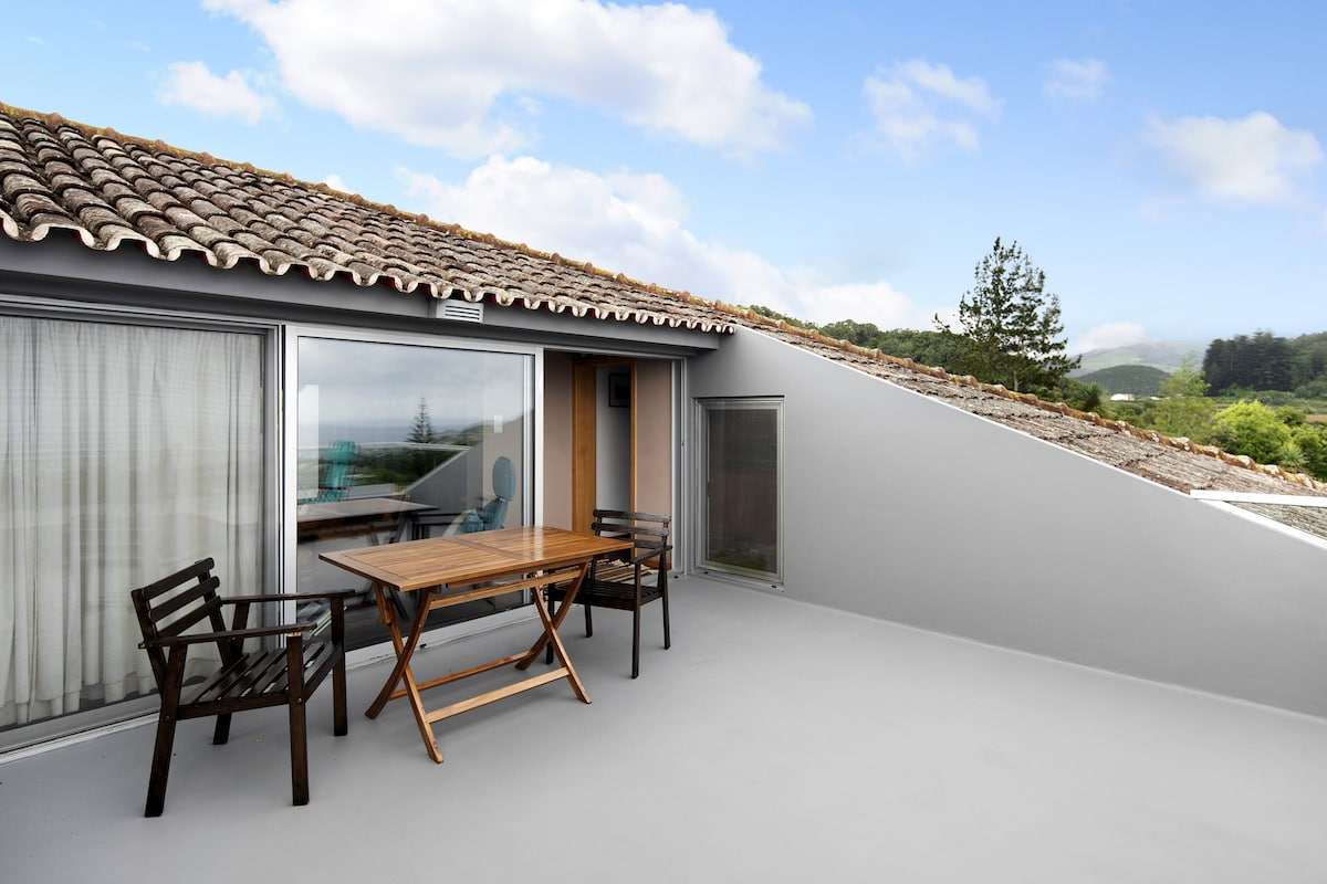 An outdoor terrace features a wooden dining table accompanied by two wooden chairs. The space overlooks scenic views and is complemented by a sloped roof and large glass windows, allowing natural light to fill the area.