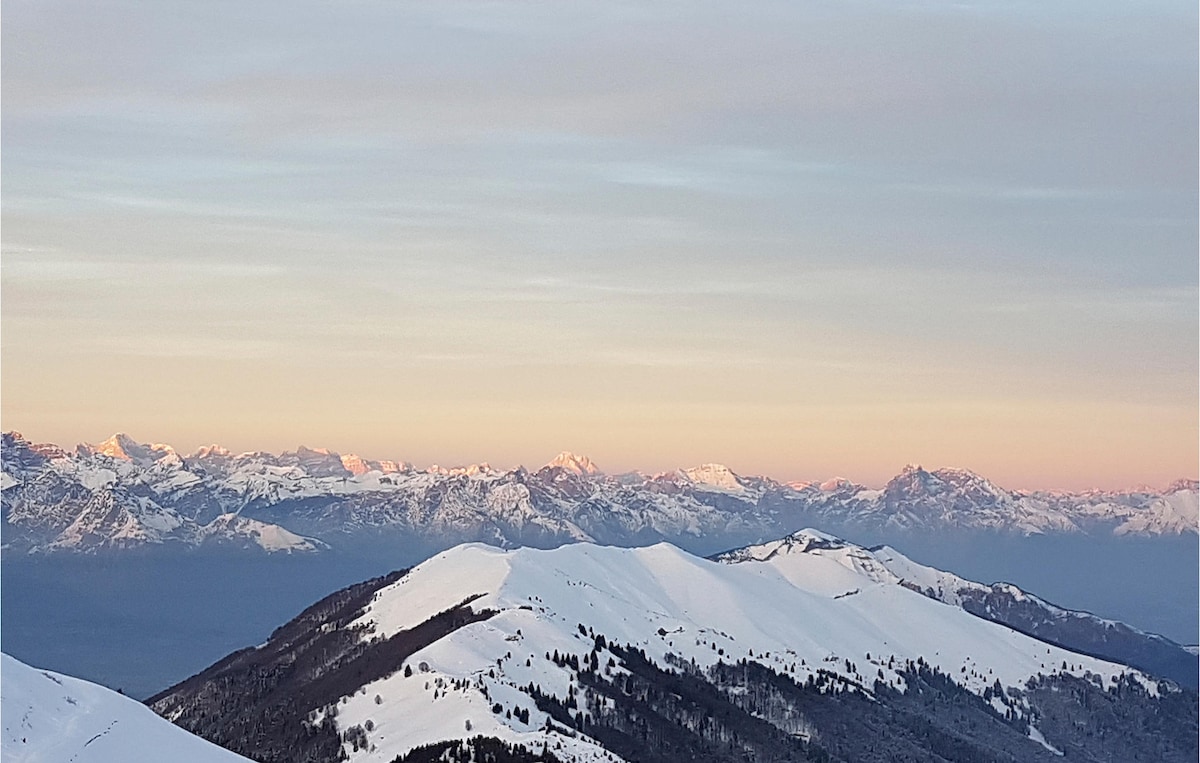 A panoramic view of snow-covered mountains stretches across the horizon, bathed in soft pastel hues of a sunrise. The rugged peaks contrast with the smooth, white slopes, creating a serene winter landscape. The vast sky reflects gentle colors, enhancing the peaceful atmosphere.