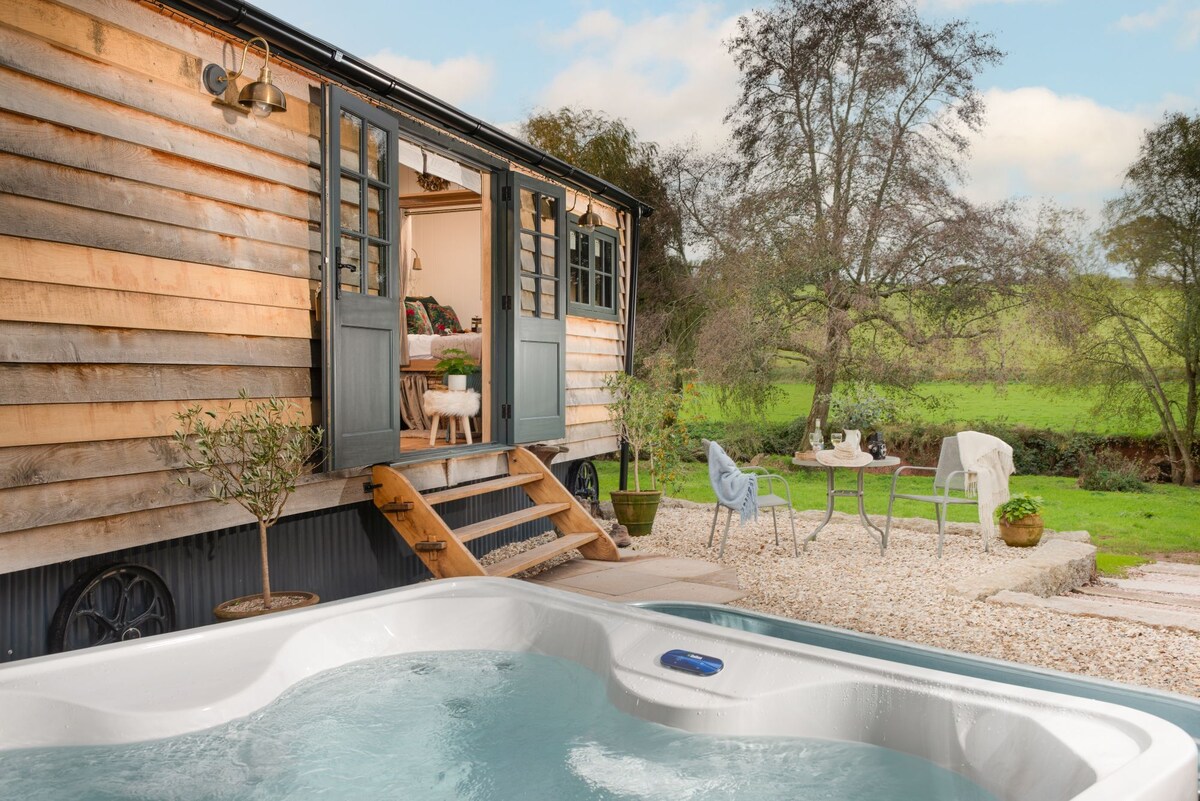 A private patio area features a hot tub, bistro table with chairs, and gravel paving, surrounded by nature. The entrance to the shepherd's hut showcases double doors opening to an inviting interior. A peaceful landscape can be seen through the greenery.