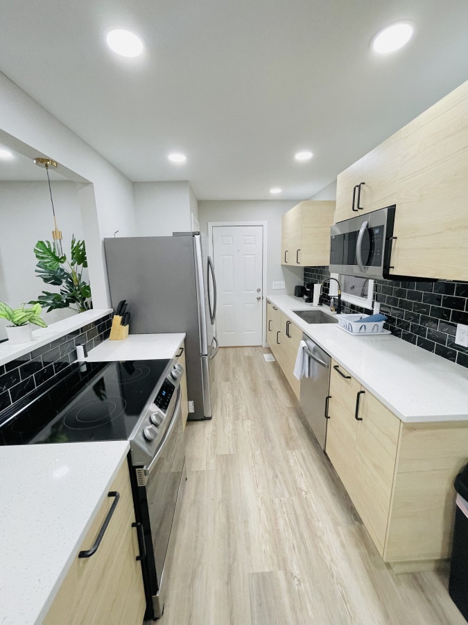 A modern kitchen features light wood cabinetry and sleek countertops. Equipped with stainless steel appliances, including a refrigerator and stove, the space is enhanced by overhead lighting. A window with a plant brings natural elements indoors, while a door provides access to the living area.