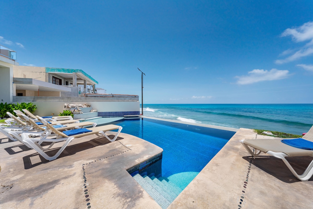 An infinity pool extends toward the horizon, offering unobstructed views of the Caribbean Sea. Comfortable lounge chairs are arranged around the pool deck. The villa structure is visible in the background, with open spaces designed for relaxation and enjoyment of the coastal scenery.