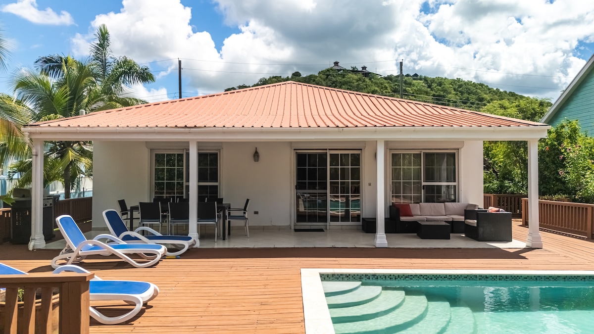 A spacious villa with a red-tiled roof is set against a backdrop of lush greenery. The outdoor area features lounge chairs and a covered seating arrangement, offering a perfect spot for relaxation. A swimming pool is visible, reflecting the blue sky and nearby palm trees.