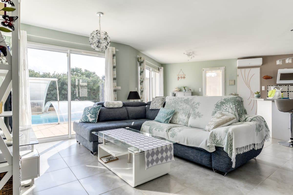 A comfortable living area features a spacious sectional sofa draped with a light throw. Natural light fills the room through large sliding glass doors, providing a view of the private pool. A simple coffee table sits in front of the sofa, with a backdrop of modern kitchen elements.