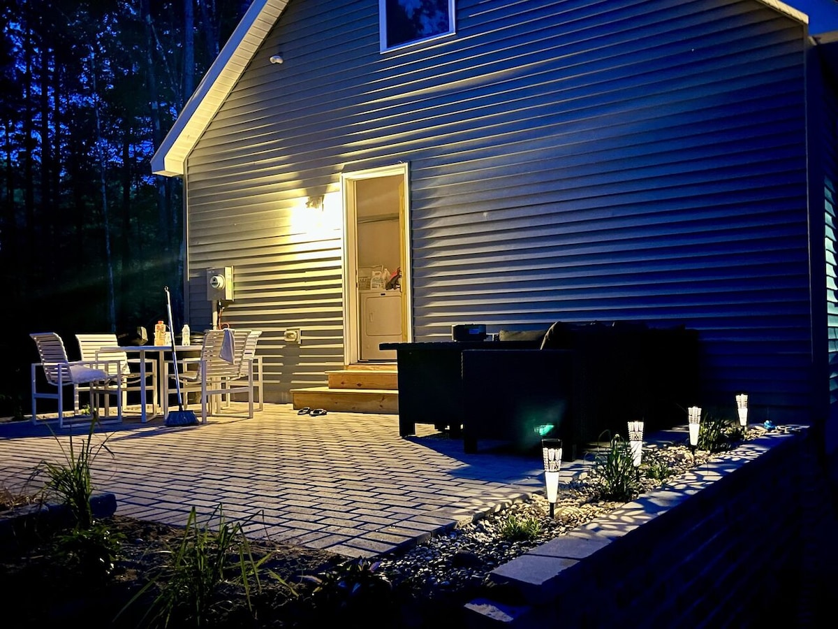 The exterior of the cottage is shown at dusk, illuminated by warm lighting. A patio area with a dining set and lounge seating is present, bordered by landscaped stones and low-lying lights. The entrance to the home is visible, leading to the interior.
