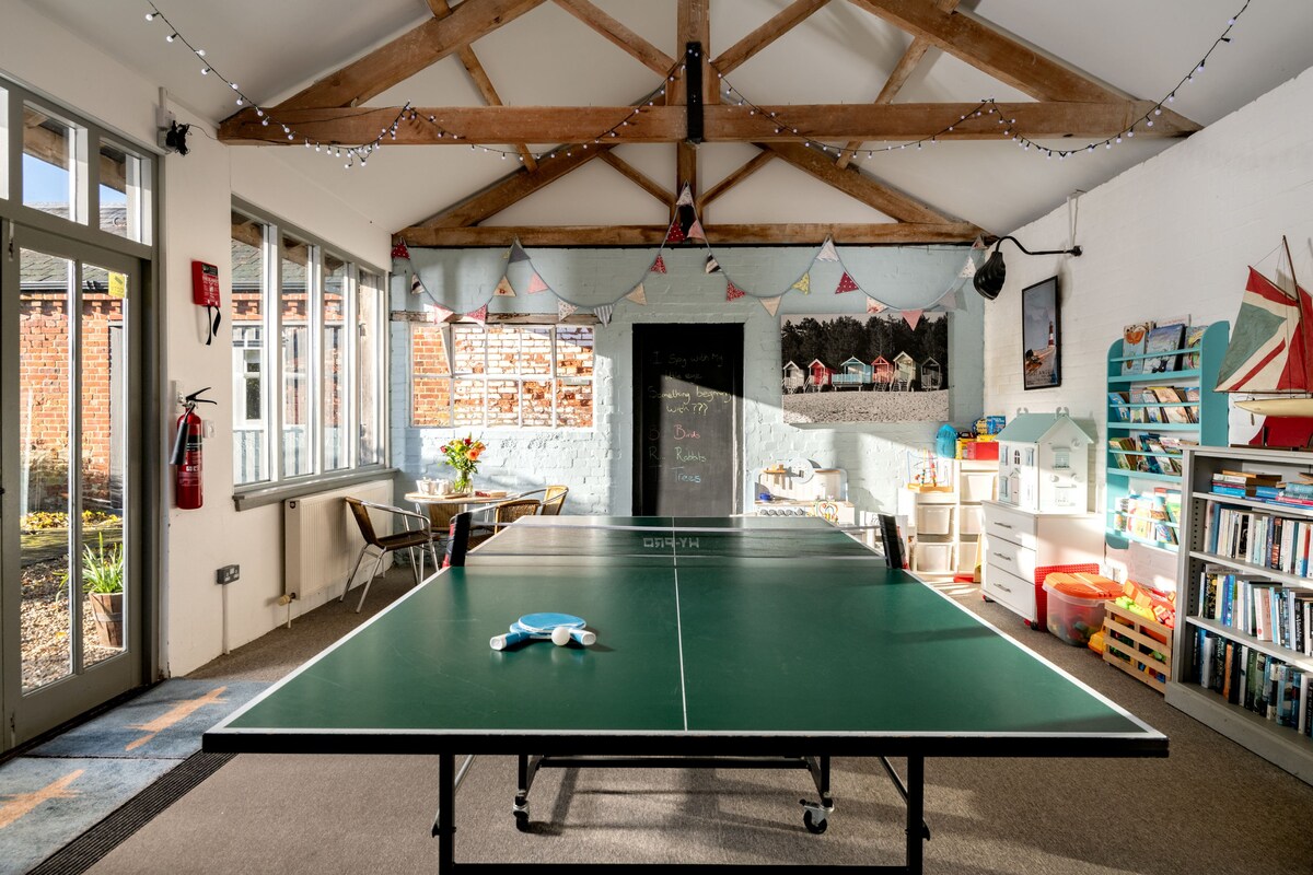 A games room is showcased with a large green ping pong table as the central focus. Features include exposed beams on the ceiling, colorful decorations on the walls, and a chalkboard area with playful drawings. Natural light fills the space through large windows, enhancing the cheerful atmosphere.